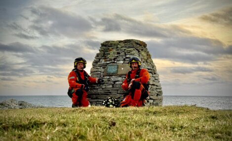 Two people in orange gear kneel by a stone monument on a grassy area near the sea, under a cloudy sky.