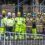 A group of construction workers and firefighters wearing high-visibility clothing and helmets stand together in front of scaffolding at a work site.