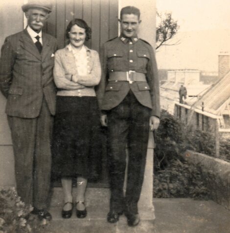 Three people, two men in suits and uniform and a woman in a cardigan and skirt, stand outdoors on steps. A greenhouse is visible in the background.