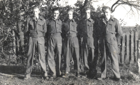 Five men in military uniforms stand in a row outdoors, in front of a fence and trees, posing for a group photo.