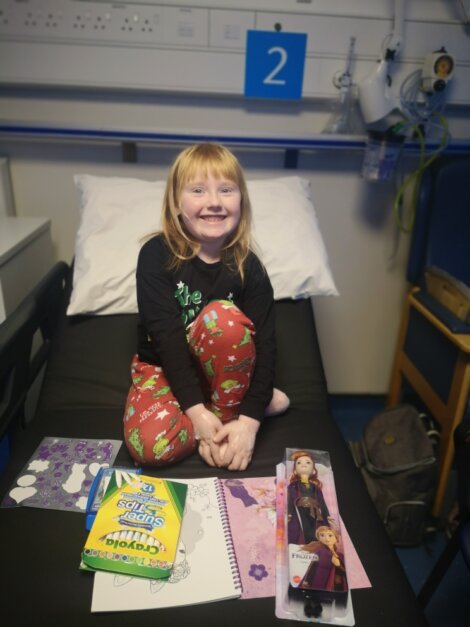A child with red hair sits on a hospital bed, smiling. Nearby are a coloring book, crayons, and a doll.