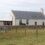 A white stone house with a gray roof sits behind a stone wall and wire fence on a grassy plot, under a cloudy sky.