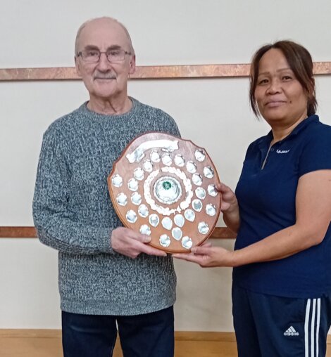 Two people standing indoors holding a wooden shield-shaped award. The man is on the left wearing a gray sweater, and the woman is on the right wearing a navy blue shirt.