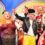 A group of people in colorful costumes perform on stage in front of a rainbow-themed backdrop. The crowd includes adults and children, with one person dressed as a police officer in front.