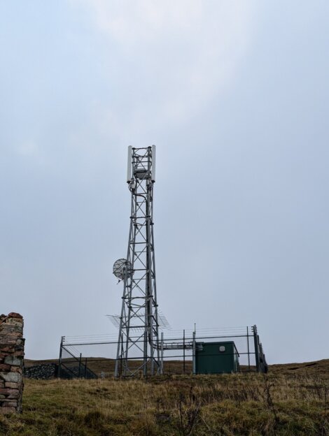 A tall metal communication tower stands on a grassy hill, enclosed by a fenced area with a small green building and a satellite dish nearby.
