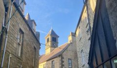 A narrow alleyway flanked by old stone buildings with a clock tower in the background under a clear blue sky.