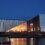 Modern waterfront building with a geometric facade illuminated from within at dusk, reflecting on the water. A small boat is moored in the foreground.