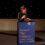 Person speaking at a podium with a sign that reads "Welcome to The Saltire Scotland’s National Book Awards.