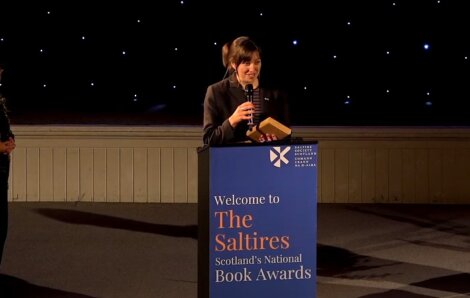 Person speaking at a podium with a sign that reads "Welcome to The Saltire Scotland’s National Book Awards.