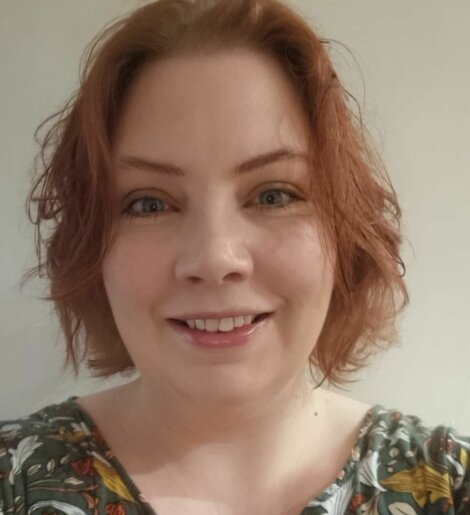 A person with short, wavy red hair is smiling at the camera. They are wearing a floral patterned top.