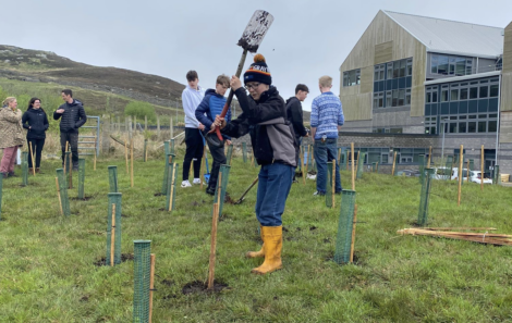 People participating in a tree planting event outside a building. A person in the foreground is digging with a shovel.