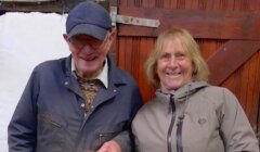 Two smiling people stand side by side in front of a wooden door. The man wears a cap and glasses, and the woman wears a hooded jacket.