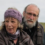 Two older adults, a woman wearing a purple headband and a man with glasses and a beard, sit outdoors with grassy fields in the background.