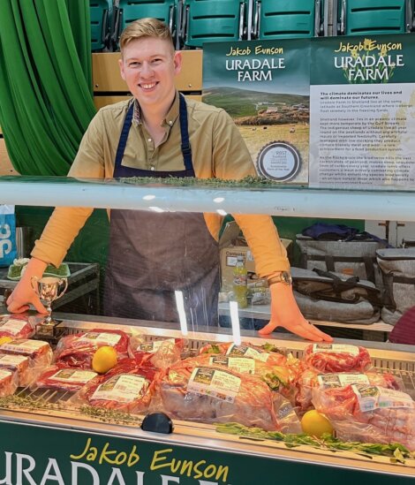 Person in an apron stands behind a display of packaged meats labeled "Jakob Eunson Uradale Farm," with information posters in the background.