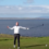A person stands on a grassy golf course holding a club, with a lake and distant hills in the background under a partly cloudy sky.