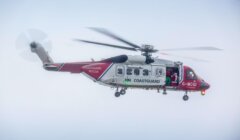 A red and white HM Coastguard helicopter in flight with visible spinning rotors and the side door open.