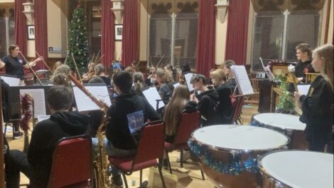 An orchestra rehearses in a hall with red curtains and a decorated Christmas tree. Musicians are seated with instruments and sheet music stands.