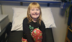A smiling child with red hair sits on a bed, wearing festive pajamas in a room with medical equipment.