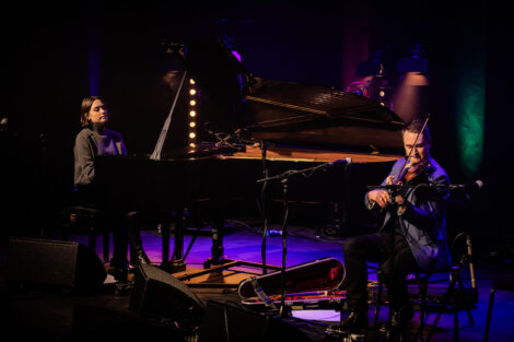 Two musicians perform on stage; one plays a grand piano, and the other plays a violin. The stage is dimly lit with colorful lights.
