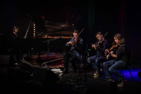 A pianist and three violinists perform on a dimly lit stage, surrounded by microphones and music equipment.