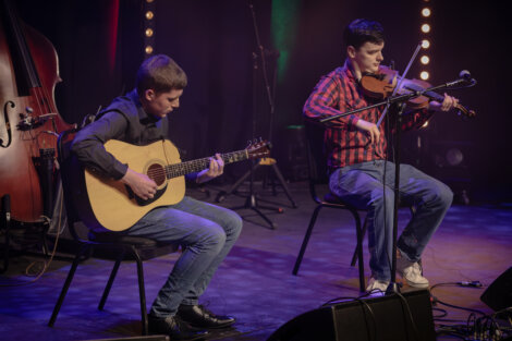 Two musicians perform on stage; one plays a guitar while the other plays a fiddle. They are seated, and there is stage lighting and a double bass in the background.