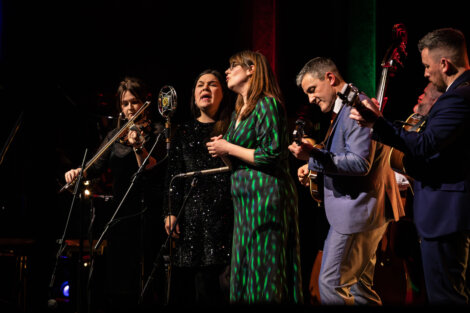 A band of musicians performs on stage with a microphone, violin, double bass, guitar, and mandolin. They are singing and playing instruments under colorful stage lights.