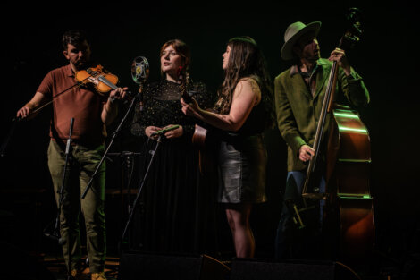 Four musicians perform on stage. The group includes a violinist, a singer, a guitarist, and a double bassist. They are focused on their performance under stage lighting.
