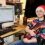 Person in a festive sweater and Santa hat holding a guitar, sitting at a desk with audio production software open on a computer screen.