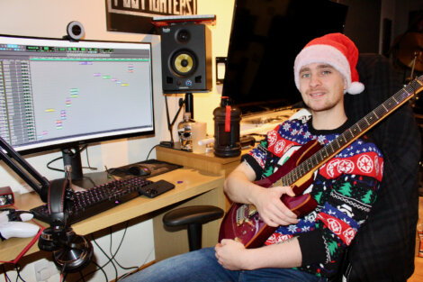 Person in a festive sweater and Santa hat holding a guitar, sitting at a desk with audio production software open on a computer screen.