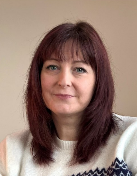 A woman with shoulder-length red hair and a white patterned sweater sits against a neutral background, looking at the camera.