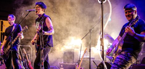 Band performing on stage with guitars and a drum set, surrounded by bright stage lights and smoke.