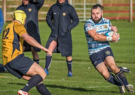A rugby player in a blue patterned jersey runs with the ball, facing an opponent in a yellow jersey. Two coaches in long coats observe from the sideline.