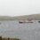 A red and white boat is escorted by two smaller tugboats in a grey, misty harbor, with hills and a few buildings in the background.