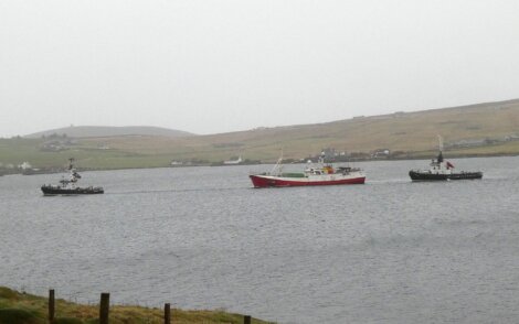 A red and white boat is escorted by two smaller tugboats in a grey, misty harbor, with hills and a few buildings in the background.