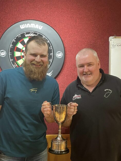 Two men holding a trophy stand in front of a dartboard, smiling.