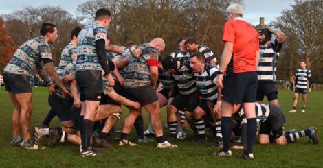 Two rugby teams in a scrum during a match on a grassy field, with a referee nearby.