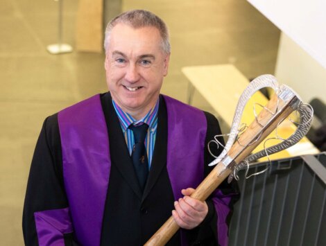 A man in a purple and black gown holds a ceremonial staff while standing on a staircase indoors.