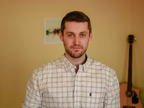 A man wearing a plaid shirt stands in a room with light-colored walls. A guitar leans against the wall in the background.