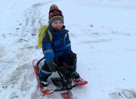 A child in a blue winter outfit and colorful hat is sitting on a red sled in the snow, smiling.
