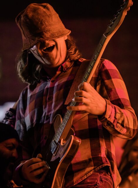 Person playing an electric guitar, wearing a plaid shirt and a bucket hat, with a blurred background.