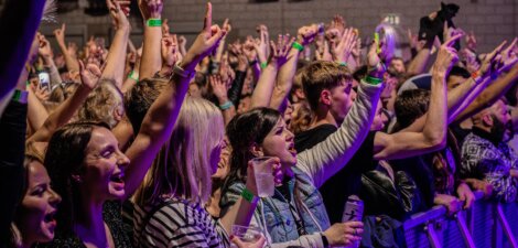 A crowd at a concert enthusiastically cheers and raises their hands near the stage, with some holding drinks and wearing wristbands.
