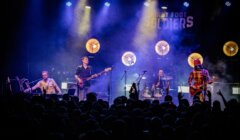 A band performs on stage with guitars, keyboard, and drums. Bright spotlights illuminate the scene, and a crowd watches from the foreground.