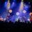 A band performs on stage with guitars, keyboard, and drums. Bright spotlights illuminate the scene, and a crowd watches from the foreground.