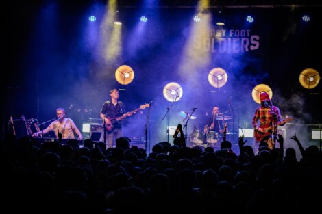 A band performs on stage with guitars, keyboard, and drums. Bright spotlights illuminate the scene, and a crowd watches from the foreground.