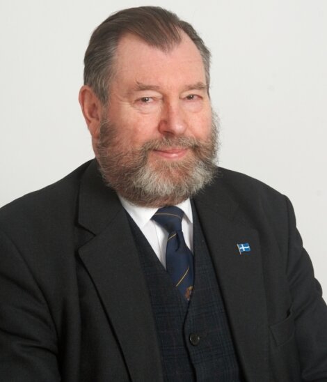 A man with a beard and mustache wearing a suit and tie poses against a plain background.