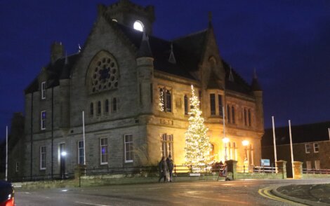 A historic stone building at night with a large, illuminated Christmas tree in front, framed by glowing windows and streetlights.