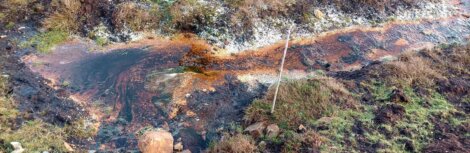 A narrow, polluted stream runs through grass and soil, showing orange and black discoloration, with a thin wooden stick placed upright nearby.