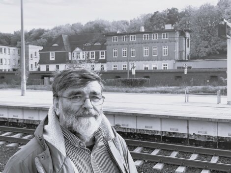 A man with glasses and a beard stands near train tracks, with buildings and trees in the background. The image is in black and white.