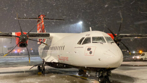 A white propeller plane is parked on a snowy tarmac at night, with snow falling and airport lights in the background.