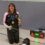 A woman kneels indoors holding a trophy and fencing sword, wearing a medal. A digital scoreboard behind her shows a score of 15:14 with a time of 3:00.